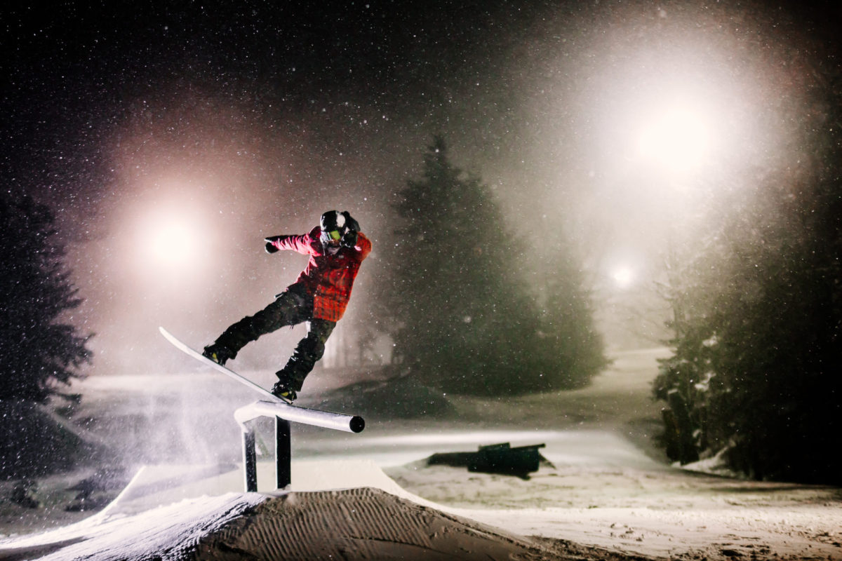 A snowboarder doing a front lipslide in a red plaid jacket at night